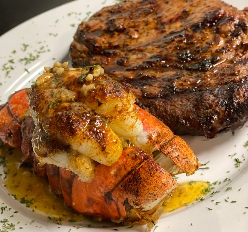 A plate featuring grilled lobster tail topped with butter and a juicy steak, garnished with herbs.