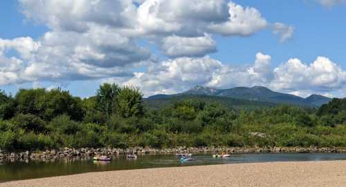 A serene river scene with kayakers, lush greenery, and mountains under a partly cloudy sky.