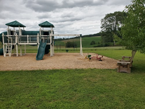 A playground with a slide and swings on gravel, surrounded by green grass and fields under a cloudy sky.