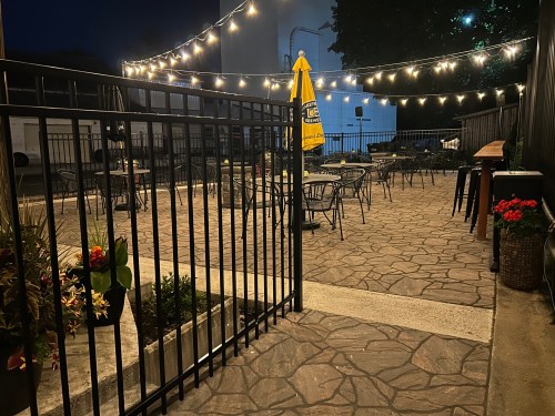 A dimly lit outdoor patio with string lights, tables, and chairs, surrounded by a black fence and potted plants.