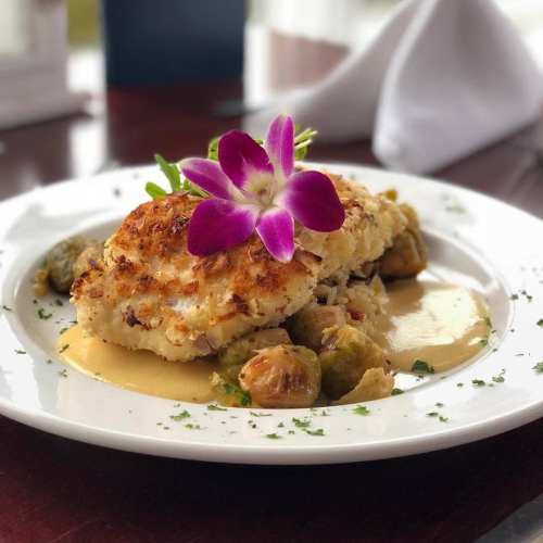A plated dish featuring crab cakes, Brussels sprouts, and a creamy sauce, garnished with a purple flower.