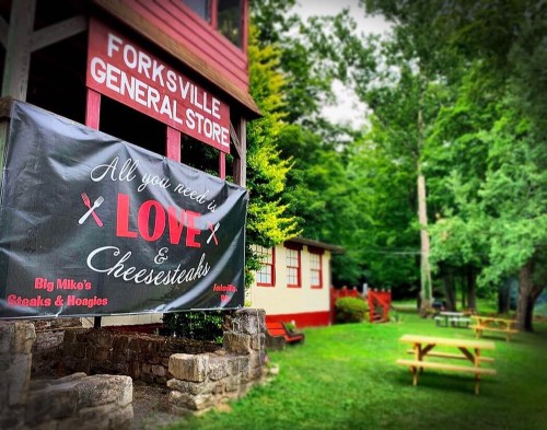 A red general store with a banner reading "All you need is LOVE & Cheesesteaks" surrounded by greenery and picnic tables.
