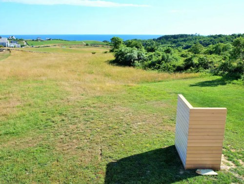 A grassy field with a wooden structure in the foreground, overlooking a blue ocean and green hills in the background.