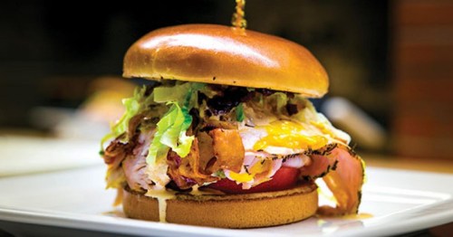 A delicious burger stacked with lettuce, tomato, bacon, and melted cheese on a toasted bun, served on a white plate.