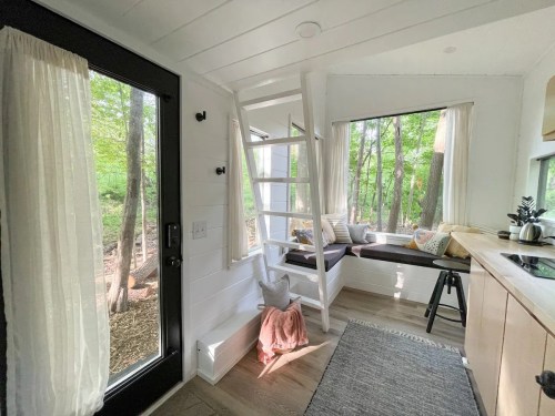 Cozy interior of a tiny house with large windows, a ladder, and a seating area surrounded by trees.