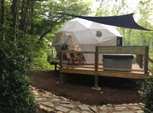 A geodesic dome cabin on a wooden deck, surrounded by trees and a stone path, with outdoor seating and a hot tub.
