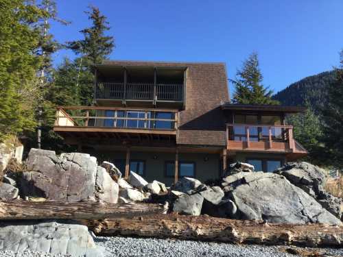 A modern house with multiple balconies, surrounded by large rocks and trees, set against a clear blue sky.