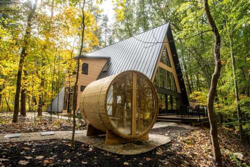 A modern A-frame cabin in a forest, featuring large windows and a wooden sauna outside, surrounded by autumn foliage.