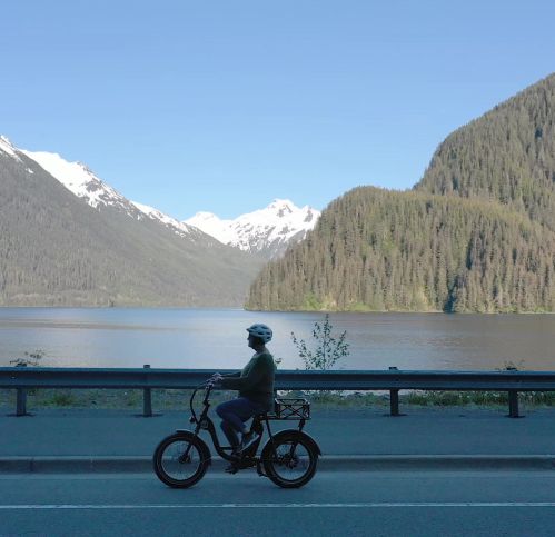 A person rides an electric bike along a scenic road by a lake, surrounded by mountains and greenery.