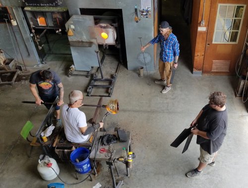 A group of four people working in a glassblowing studio, shaping molten glass and using various tools.