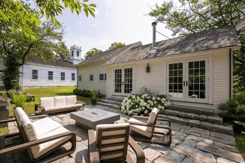 A cozy outdoor patio with modern furniture and a fire pit, surrounded by greenery and a charming white house.