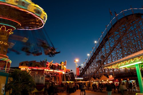 A vibrant amusement park at dusk, featuring a spinning swing ride and a towering wooden roller coaster.