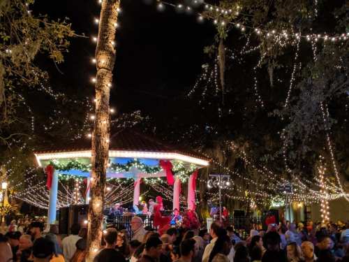 A festive night scene with a crowd gathered around a decorated gazebo, illuminated by string lights and holiday decorations.