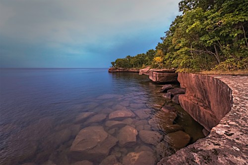 A serene lakeside view with rocky cliffs and lush greenery under a moody sky.