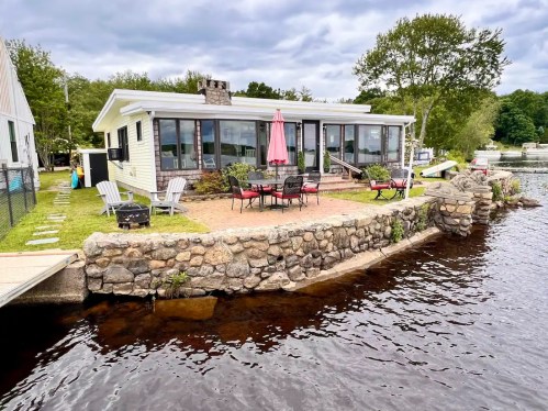 A lakeside house with a stone patio, surrounded by chairs and a pink umbrella, under a cloudy sky.