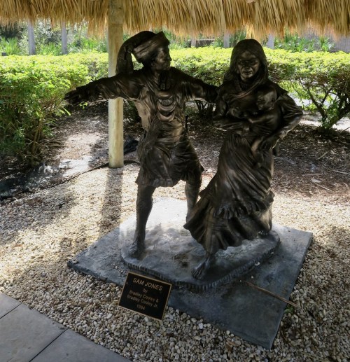 Bronze statue of a man and woman dancing, with a plaque reading "Sam Jones" in a tropical setting.