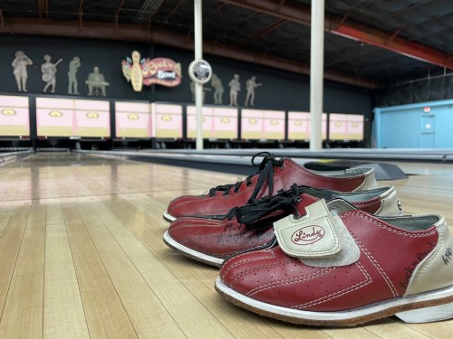 A pair of red bowling shoes on a wooden lane, with a retro bowling alley backdrop featuring silhouettes and neon signs.
