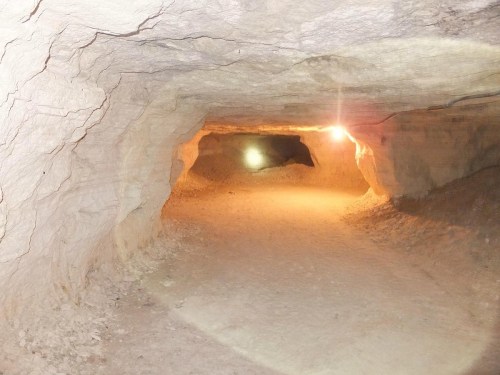 A dimly lit cave interior with smooth, light-colored walls and a sandy floor, featuring a pathway leading deeper inside.