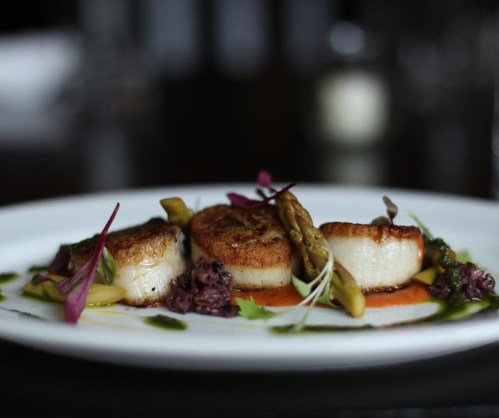 Three seared scallops on a plate, garnished with asparagus, colorful herbs, and a drizzle of sauce.