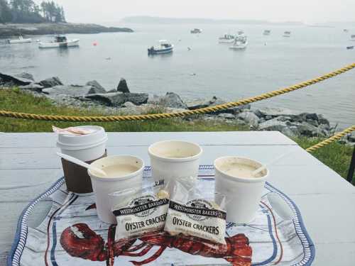 Three cups of clam chowder and oyster crackers on a table by the water, with boats in the background.