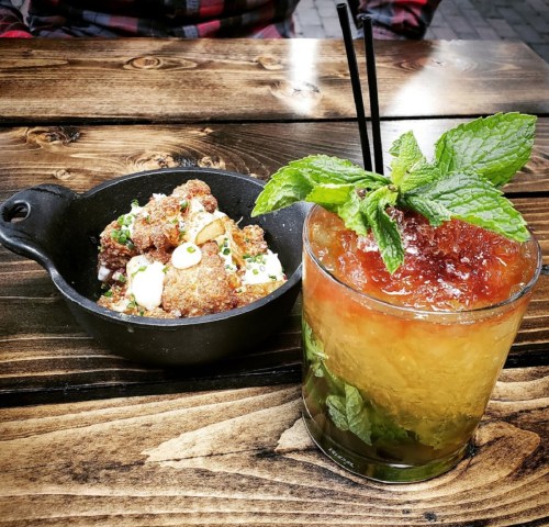 A refreshing cocktail with mint sits beside a small skillet of crispy bites on a wooden table.