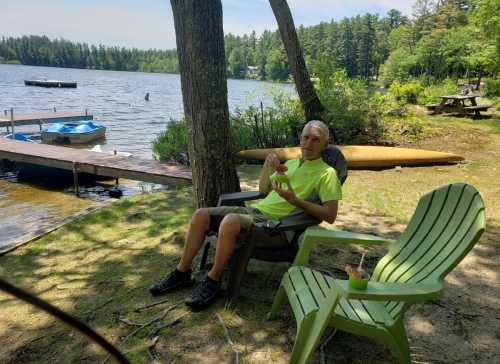 A person in a bright shirt sits in a chair by a lake, enjoying a snack with boats and trees in the background.