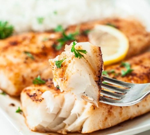 A fork holding a piece of seasoned fish, garnished with parsley, alongside a plate of rice and lemon slices.