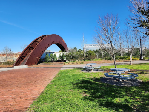 A curved metal arch in a park with picnic tables and trees under a clear blue sky.