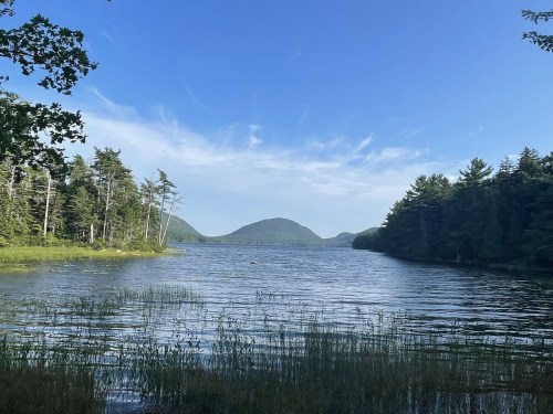 A serene lake surrounded by trees, with a distant mountain under a clear blue sky and wispy clouds.