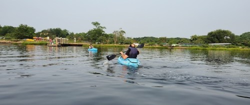 Two people kayaking on a calm waterway, with greenery and a dock in the background.