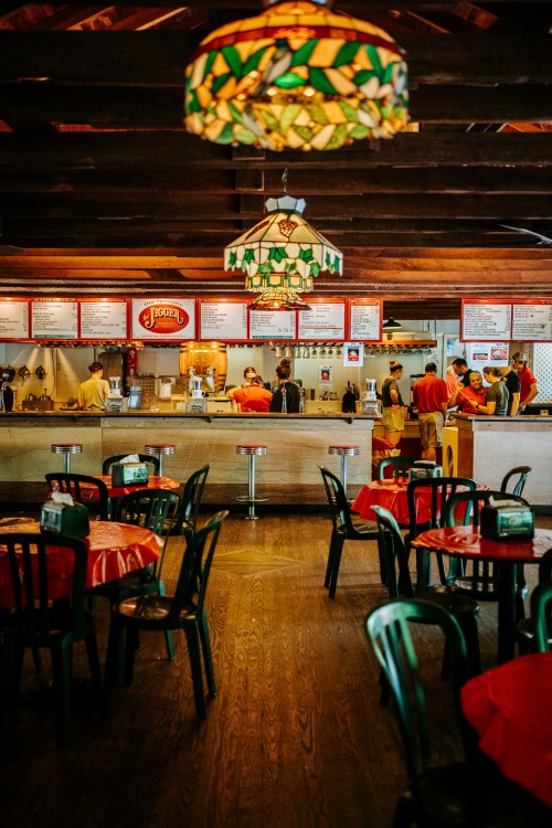 Interior of a casual restaurant with colorful stained glass lamps, red tablecloths, and a busy counter area.