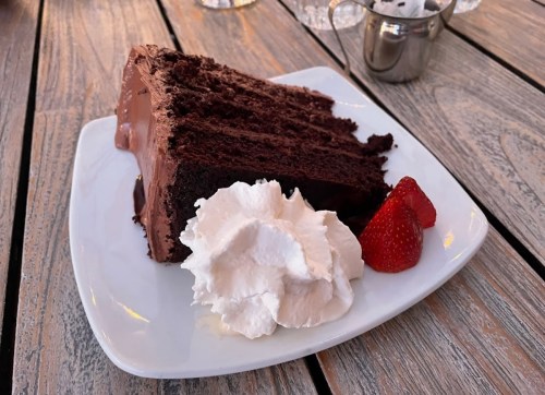 A slice of chocolate cake with rich frosting, served with whipped cream and a fresh strawberry on a white plate.