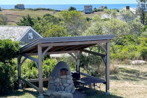 A stone outdoor oven under a wooden gazebo, with a view of the ocean and distant houses in the background.