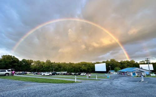 A vibrant rainbow arcs over a drive-in theater surrounded by trees and parked cars under a cloudy sky.