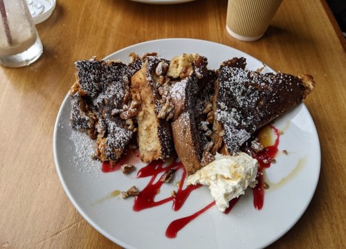 A plate of French toast topped with powdered sugar, nuts, and a dollop of whipped cream, drizzled with sauce.