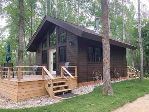 A cozy brown cabin with a wooden deck, surrounded by trees and featuring a stone pathway and decorative wheels.