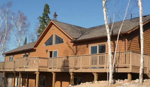 A wooden house with a large deck, surrounded by trees and a clear blue sky.