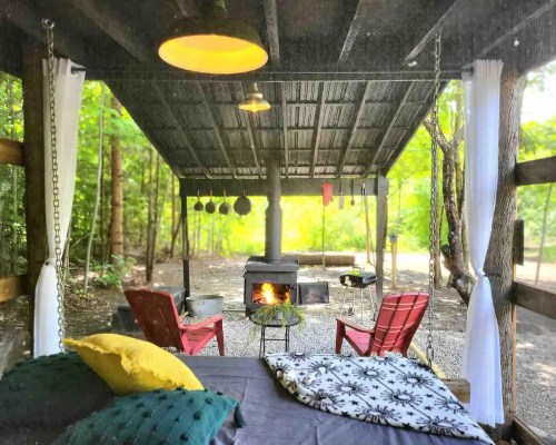 Cozy outdoor space with a fire pit, two chairs, and a bed, surrounded by trees and natural light.