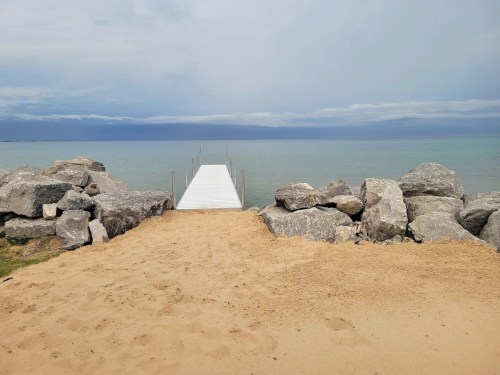 A sandy beach leads to a wooden dock extending over calm water, surrounded by large rocks under a cloudy sky.