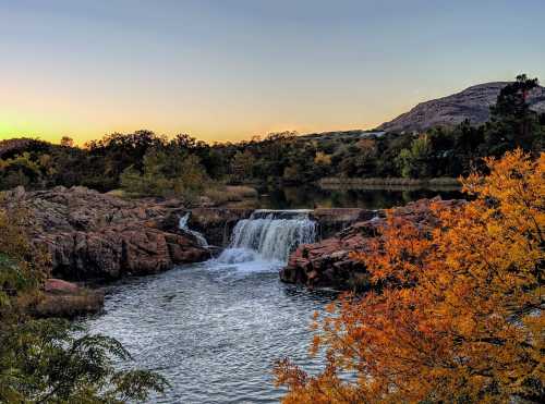 A serene waterfall cascades over rocks, surrounded by autumn foliage and a tranquil lake at sunset.