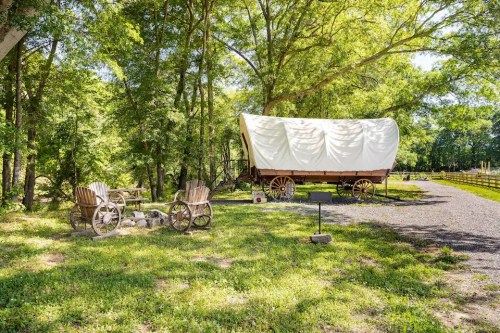 A vintage wagon with a white cover sits among trees, with two wooden chairs nearby on a grassy area.
