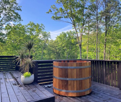 A wooden hot tub on a deck surrounded by lush green trees under a clear blue sky.