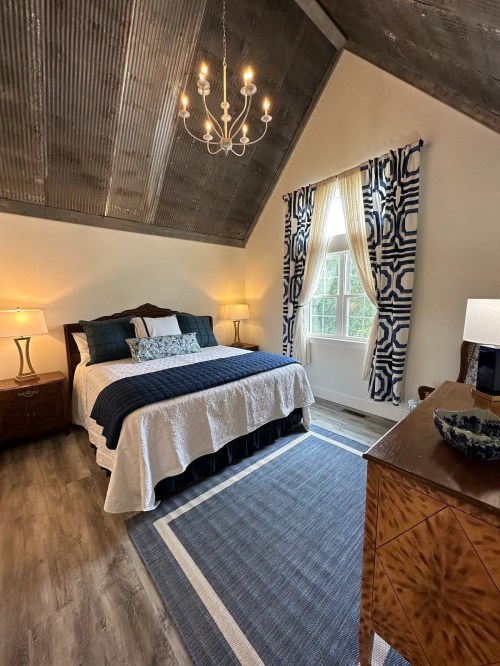Cozy bedroom with a chandelier, patterned curtains, a large bed, and wooden furniture against a rustic ceiling.