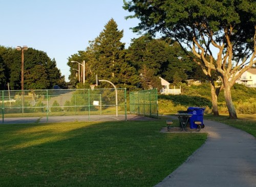 A quiet park scene featuring a basketball court, green grass, trees, and a blue recycling bin along a paved path.