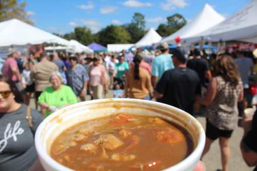 A hand holds a bowl of stew in the foreground, with a busy outdoor festival and crowd in the background.