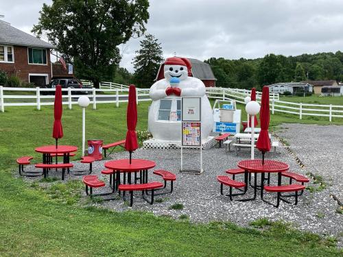 A whimsical ice cream stand shaped like a snowman, surrounded by red picnic tables and umbrellas on a grassy area.