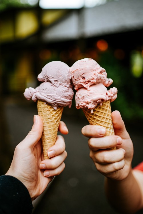 Two hands hold ice cream cones with pink and purple scoops, clinking them together in a cheerful gesture.
