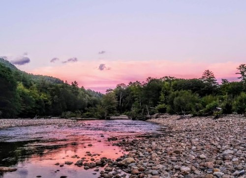 A serene river scene at sunset, with colorful skies reflecting on the water and rocky shores surrounded by trees.