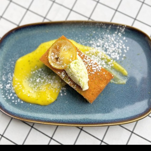 A slice of cake topped with cream and lemon, served on a blue plate with a drizzle of yellow sauce and powdered sugar.