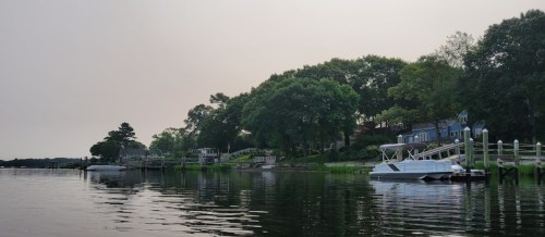 A calm river scene with boats docked, lush greenery, and houses along the shore under a hazy sky.